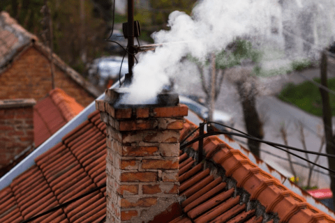 Smoke billows from a brick chimney on a rooftop with red tiles, against a backdrop of treetops.